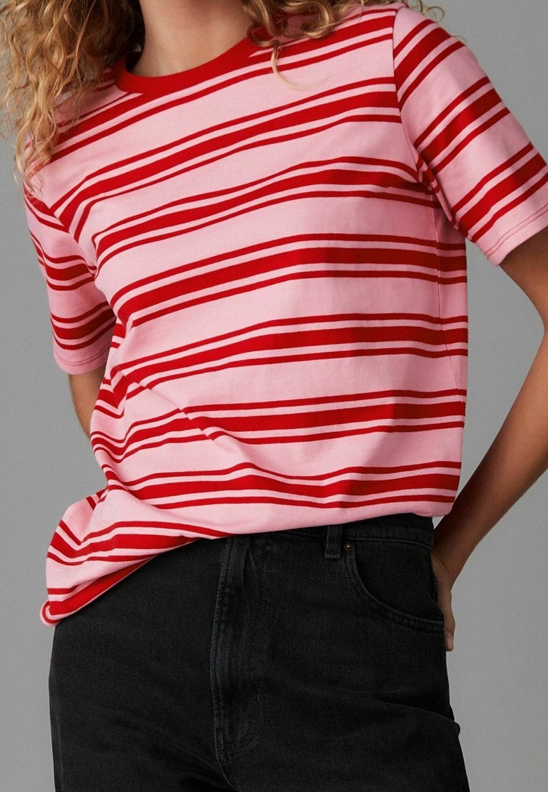 Personne portant une chemise rose à manches courtes avec des rayures horizontales rouges irrégulières, légèrement rentrée dans un jean noir taille haute.