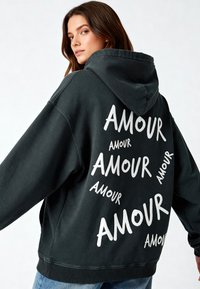 Felpa nera oversize con testo bianco "AMOUR" in diverse dimensioni e orientamenti. Tessuto morbido con tasca a marsupio e cappuccio regolabile con cordini.
