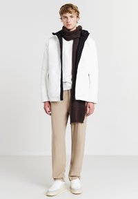 Weiße gerippte Jacke mit schwarzen Akzenten, beige Hose und einem dunkelbraunen Strickschal. Das Model trägt weiße Sneakers. Einfaches, modernes Design.