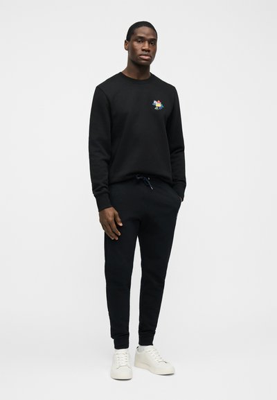 Άνδρας στέκεται με τα χέρια στις τσέπες, φοράει μαύρο φούτερ με μικρό πολύχρωμο ζωάκι, μαύρα παντελόνια φόρμας και λευκά αθλητικά παπούτσια.