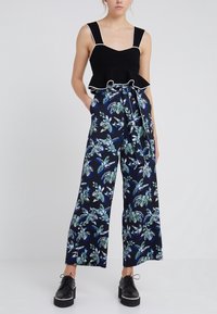 Svart peplumtop med vit kant i kombination med vida byxor med tropiskt bladmönster i grönt och blått på en mörk bakgrund.