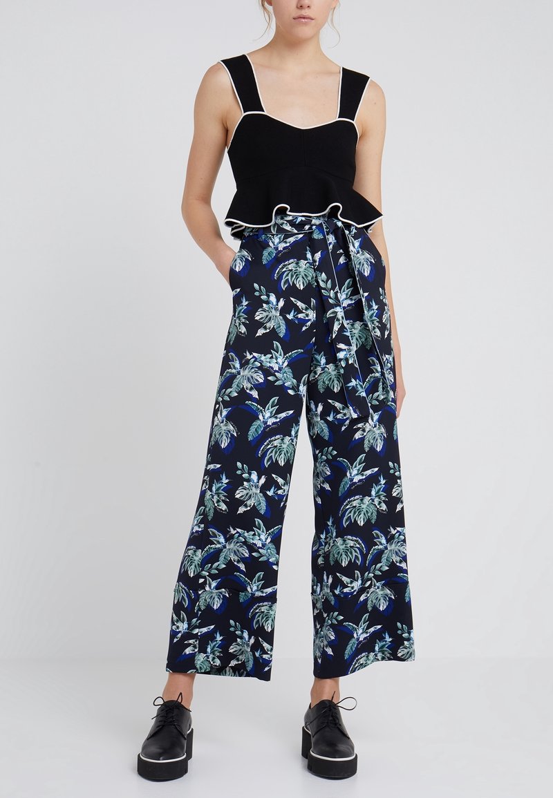 Svart peplumtop med vit kant i kombination med vida byxor med tropiskt bladmönster i grönt och blått på en mörk bakgrund.