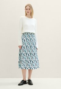 Frau in einem weißen Langarm-Pullover, einem blau-weißen, floral gemusterten, plissierten Midi-Rock und schwarzen Schnürschuhen, die vor einem einfarbigen Hintergrund steht.