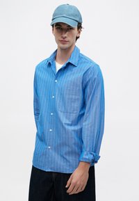 Junger Mann in einem hellblauen gestreiften Hemd mit Knöpfen, weißem T-Shirt, Jeans und einer blauen Baseballkappe, der vor einem weißen Hintergrund steht.