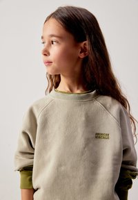 Jeune fille aux longs cheveux bruns portant un sweat-shirt beige "American Vintage", regardant vers sa gauche sur un fond clair uni.