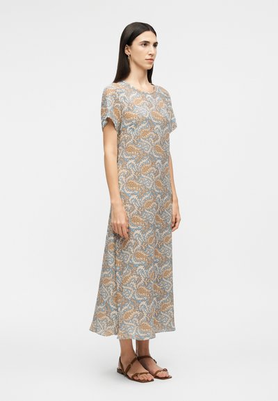 Γυναίκα που στέκεται πλαγίως φορώντας μια μίντι φόρεμα με λουλουδάτο σχέδιο σε ανοιχτό μπλε και καφέ αποχρώσεις, με κοντά μανίκια και καφέ σανδάλια.