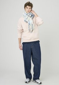 Sweatshirt rose clair, chemise marine superposée en dessous, pantalon en denim sombre ample, et écharpe rayée multicolore, le tout associé à des chaussures de sport.