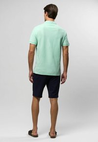 Világos zöld rövidujjú póló, tengerészkék rövidnadrággal párosítva. Az anyaga simának tűnik, és a gallér struktúrált. Barna szandálok.