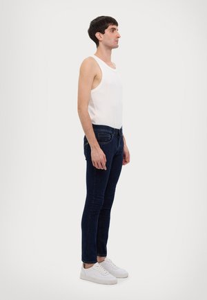 Canotta bianca senza maniche, vestibilità media, abbinata a jeans skinny in denim scuro, frontale piatto, con dettagli minimi, e sneakers bianche.