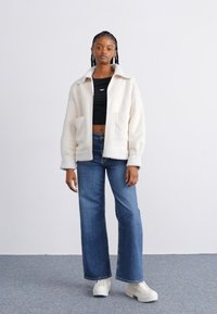 Veste en polaire couleur crème avec col, deux poches avant, portée sur un haut noir, associée à un jean bleu à jambes larges et des baskets blanches.