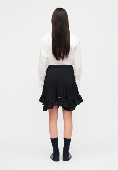 Μαύρη πλισέ φούστα με βολάν στο τελείωμα και λεπτομέρεια από δαντέλα, σε συνδυασμό με λευκό πουκάμισο με κουμπιά, μπλε κάλτσες μέχρι το γόνατο και μαύρα παπούτσια.