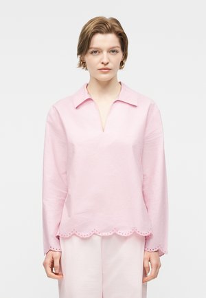 Fiatal nő rövid hajjal, világos rózsaszín galléros blúzt visel, amelyen hullámos, lyukacsos szegélyek vannak, valamint hozzáillő világos rózsaszín nadrágot.