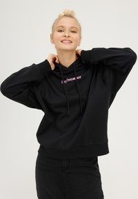 Femme souriante ajustant un sweat à capuche noir avec le texte rose "DayDreamer", portant un pantalon noir, debout devant un fond clair uni.