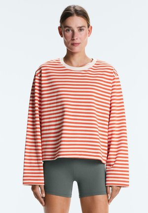 Žena stojí čelom vpred, oblečená v voľnom oranžovo-bielom pruhovanom tričku s dlhým rukávom a priliehavých tmavosivých krátkych nohaviciach na jednofarebnom pozadí.