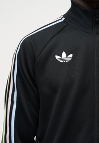 Sort lynlåstrøje med høj krave, der har pastelblå, pink og gule striber langs skulderen og et hvidt Adidas Trefoil-logo på brystet.