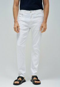 Pantalones de algodón blancos con un corte ajustado, con passadores para cinturón y bolsillos delanteros, combinados con sandalias negras y una parte superior oscura.