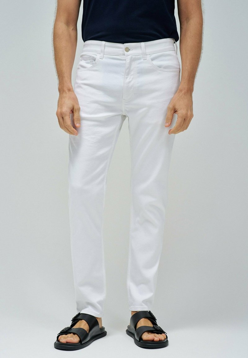 Pantalones de algodón blancos con un corte ajustado, con passadores para cinturón y bolsillos delanteros, combinados con sandalias negras y una parte superior oscura.