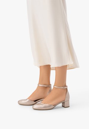 Piedi che indossano scarpe metallizzate con tacco basso e cinturini alla caviglia, abbinate a una gonna midi fluida color crema su uno sfondo bianco.