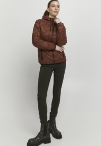Veste matelassée marron avec capuche, dotée de fermetures éclair noires, assortie à un pantalon skinny vert foncé et des bottines noires épaisses.
