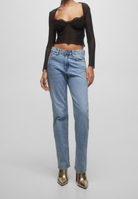 Haut noir transparent à manches longues avec bordure en dentelle, associé à un jean droit bleu clair. Le mannequin porte des escarpins dorés métalliques à bout pointu.