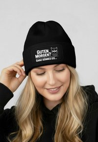 Black knit beanie with a rectangular patch featuring a white owl graphic and text: "Guten Morgen? Ganz dünnes Eis..." in bold font.