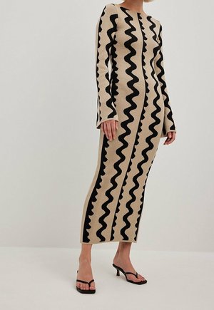 Femme portant une longue robe beige avec des rayures noires ondulées verticales, manches longues, et des sandales à talons noires ouvertes à l'avant.