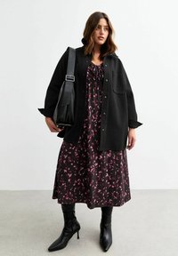 Camisa chaqueta negra de gran tamaño sobre un vestido oscuro con patrones abstractos en rosa; botas altas negras puntiagudas hasta la rodilla; gran bolso negro de hombro.