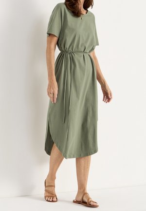 Kvinde iført olivengrøn midi kjole med korte ærmer og taljebånd, parret med tan flade sandaler, står mod en hvid baggrund.