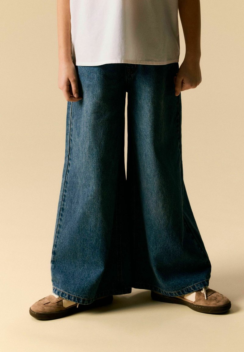 Personne portant un jean bleu à jambes larges, des baskets beiges et une chemise blanche, debout devant un fond beige uni.