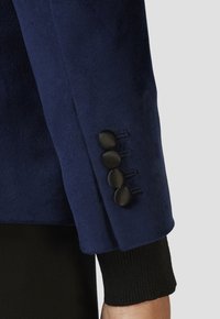 Rukáv námořnického modrého blejzru z jemného materiálu má čtyři černé knoflíky a na konci se nachází žebrované černé manžety pro kontrast a detail.