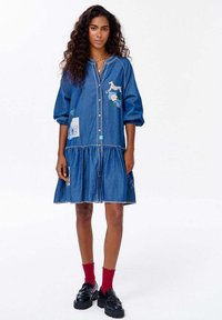 Džínová šaty s predným zapínaním na gombíky, krátkymi puffovými rukávmi a vrstveným lemom. Obsahuje výšivku a nášivky v modrej a bielej farbe. Nosené s čiernymi topánkami a červenými ponožkami.