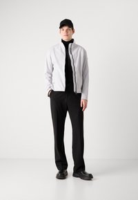 Λευκή ελαφριά ζακέτα με ψηλό γιακά, κλείσιμο με φερμουάρ και μαύρες λεπτομέρειες, συνδυασμένη με μαύρο πουλόβερ τύπου φούστα και παντελόνι, και μαύρα παπούτσια.