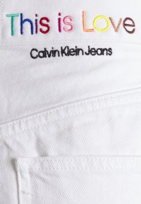 Valkoinen kangas, jossa on kirjailtu teksti "Tämä on Rakkautta" monivärisillä langoilla ja "Calvin Klein Jeans" mustalla. Sisältää taskupistot.