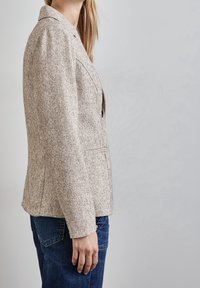 Blazer texturé beige clair avec un col cranté et deux poches avant, porté sur un jean bleu foncé. Le blazer a une coupe cintrée.