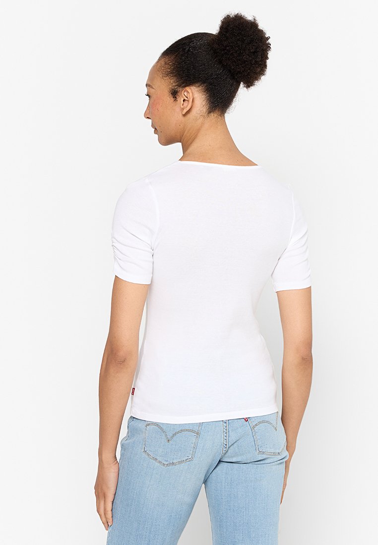 Camiseta blanca de manga corta con cuello redondo y diseño ajustado, fabricada en tela suave. Costuras visibles y detalle de fruncido lateral.