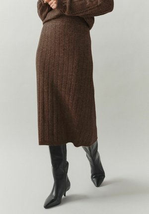 Midi-length brown ribbed knit skirt worn with black pointed-toe heeled boots, hands clasped at waist, neutral background.