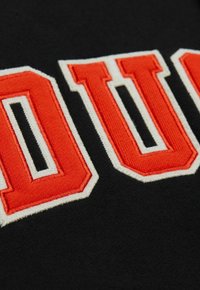 Nahaufnahme von schwarzem Stoff mit rot-weißen gestickten Buchstaben "DU" in fetter, collegeartiger Schriftart.