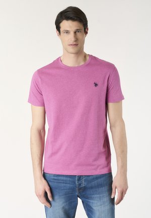 Uomo che indossa una t-shirt rosa semplice a maniche corte con un piccolo logo scuro sul petto e jeans blu, in piedi su uno sfondo neutro.