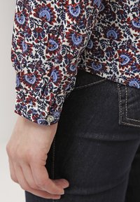 Langsleeve blouse met een witte stof met een bloemenpatroon in rood, blauw en zwart, afgewerkt met een knoopmanchet en een aansluitende snit.