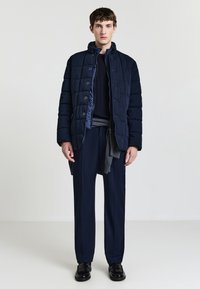 Giacca puffer trapuntata blu navy con colletto alto, caratterizzata da chiusura con bottoni, indossata sopra un maglione scuro e pantaloni su misura. Le scarpe nere completano l'outfit.
