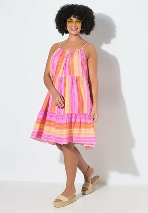 Lachende vrouw met krullend haar die gele zonnebrillen, een roze gebloemde mouwloze jurk en beige plateauzandalen draagt.