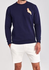 Felpa blu navy con maniche lunghe, collo tondo e un ricamo testurizzato di un uccello color crema sul petto. Abbinata a pantaloni corti bianchi.