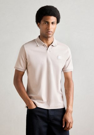 Beige poloshirt met een kraag, korte mouwen en een klein logo op de borst. Draagt met een donkere short. Soepele textuur.
