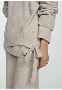 Béžový pletený sveter s prekrývajúcim sa dizajnom, rebrovanými manžetami a detailom viazania na boku. Mäkká textúra s jemným leskom.