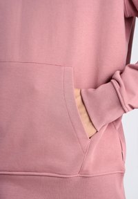Sudadera rosa con un bolsillo frontal, puños de canalé y una textura suave. Se pueden ver detalles de costura a lo largo de los bordes y el bolsillo.