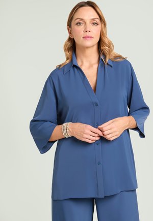 Femme aux cheveux blonds ondulés portant une blouse bleue ample à boutons et un pantalon assorti, ajustant un bracelet au poignet, fond neutre.