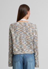 Žena s dlouhými hnědými vlasy, oblečená v barevném pleteném svetru a modrých džínech, stojící zády k jednobarevnému šedému pozadí.