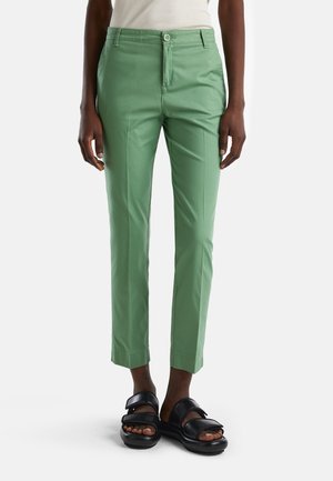 Pantalones verdes de ajuste delgado combinados con sandalias negras de punta abierta en una persona sobre un fondo claro y liso.