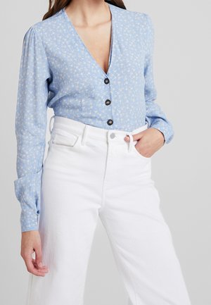 Modrá květovaná halenka s hlubokým výstřihem do V, zapínáním na knoflíky a nabíranými rukávy, v kombinaci s bílými kalhotami s vysokým pasem. Hladká textura látky.