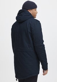 Parka de culoare albastru marin, cu glugă atașată și buzunare laterale, având o textură netedă și o talie ajustabilă pentru o potrivire perfectă.